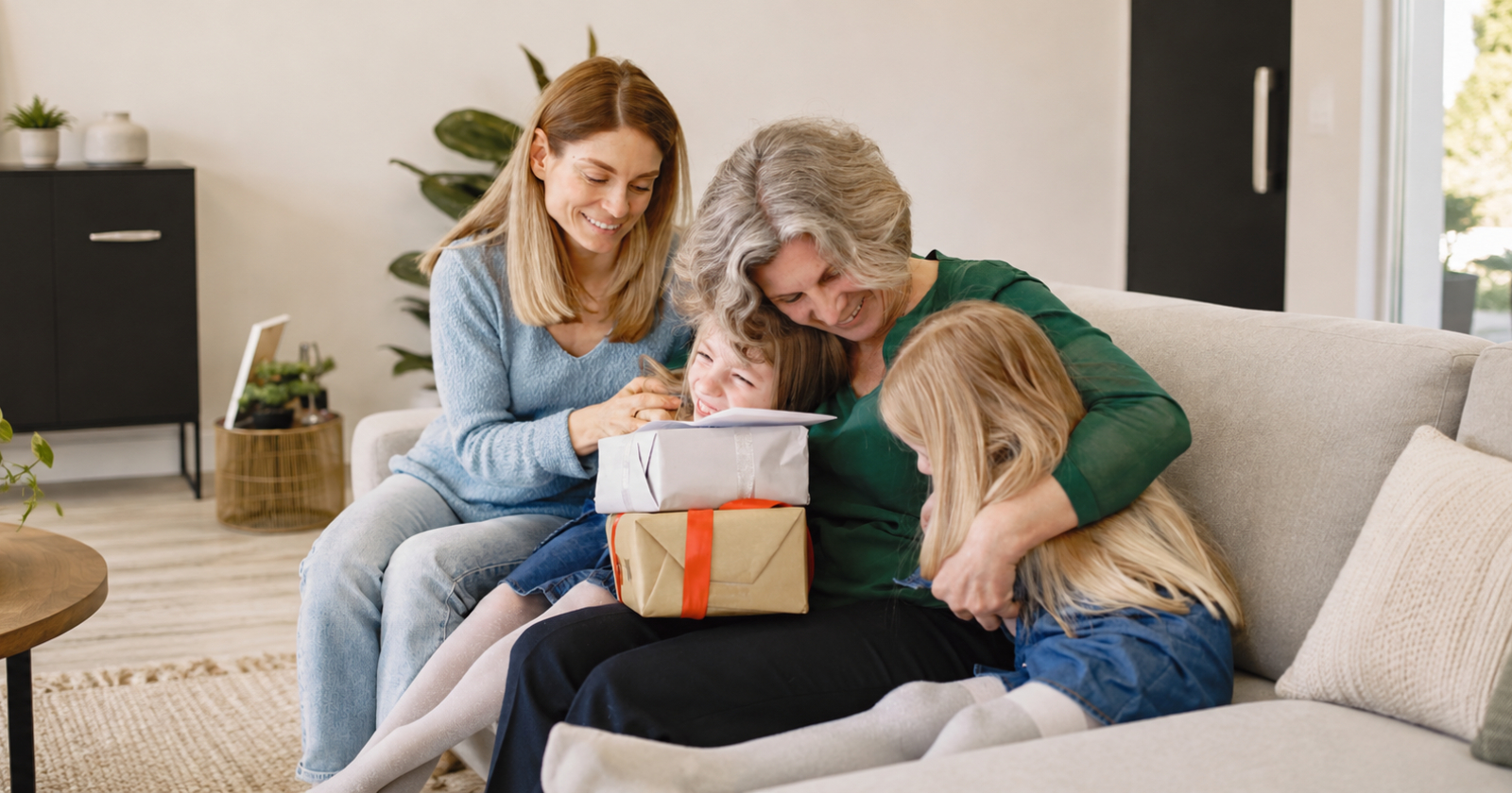 A grandmother hugs her granddaughters on a sofa beside wrapped gifts as their mother looks on.