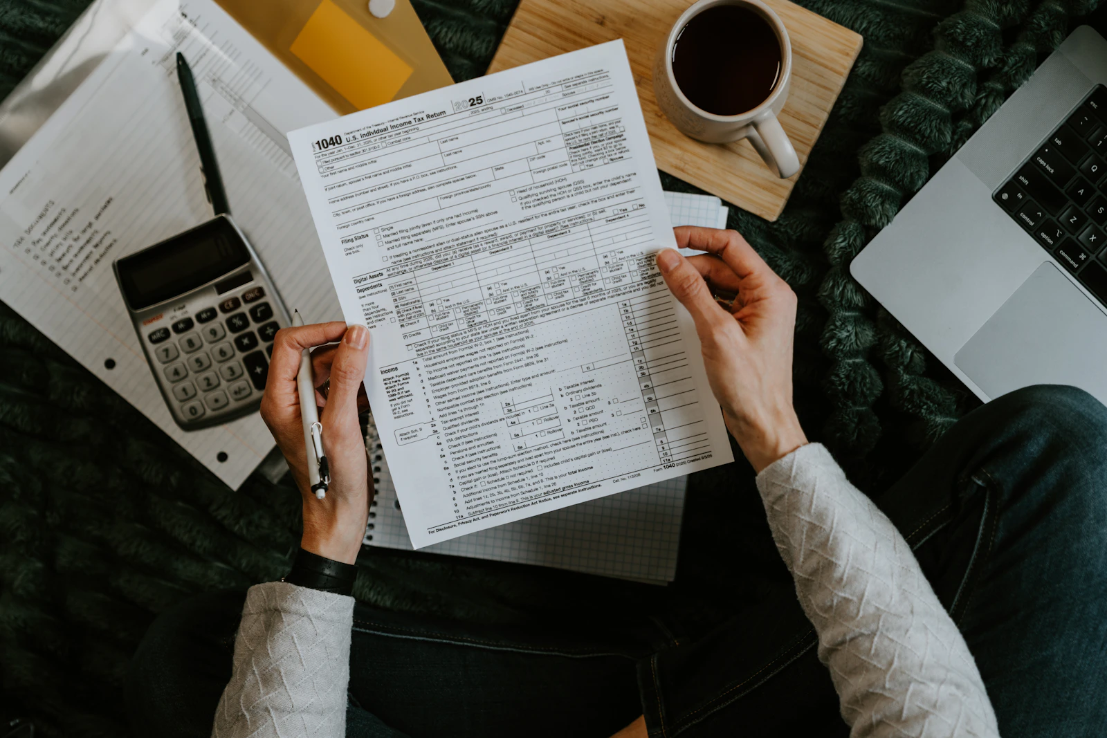 Hands holding tax forms with a calculator and laptop on a wooden desk.
